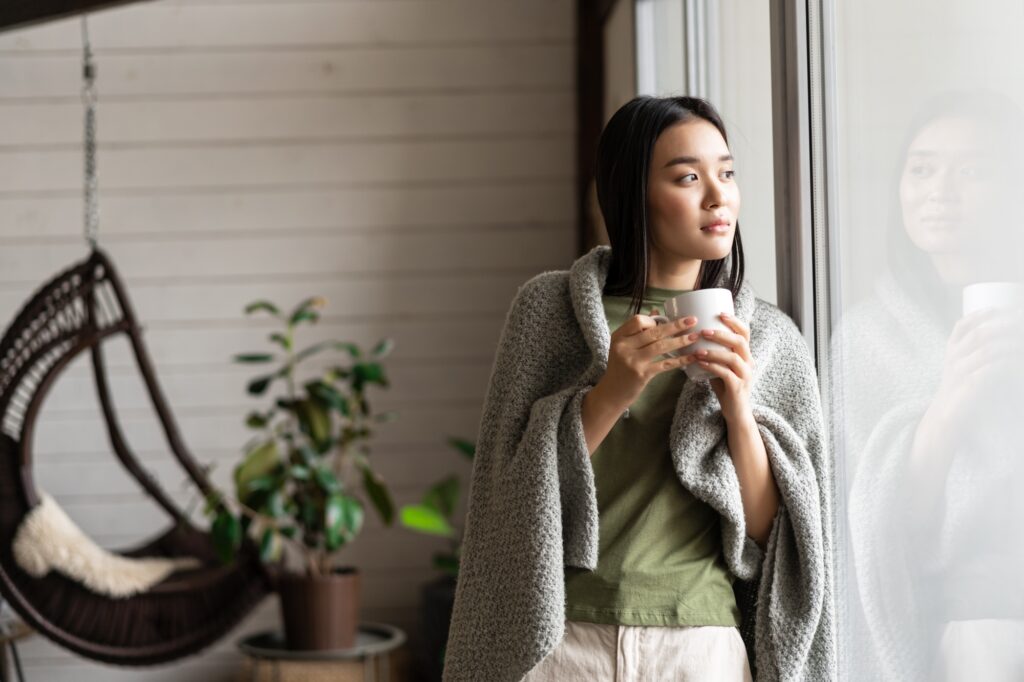 Beautiful asian woman wrapped in blanket, leaning on window and looking outside, drinking hot tea