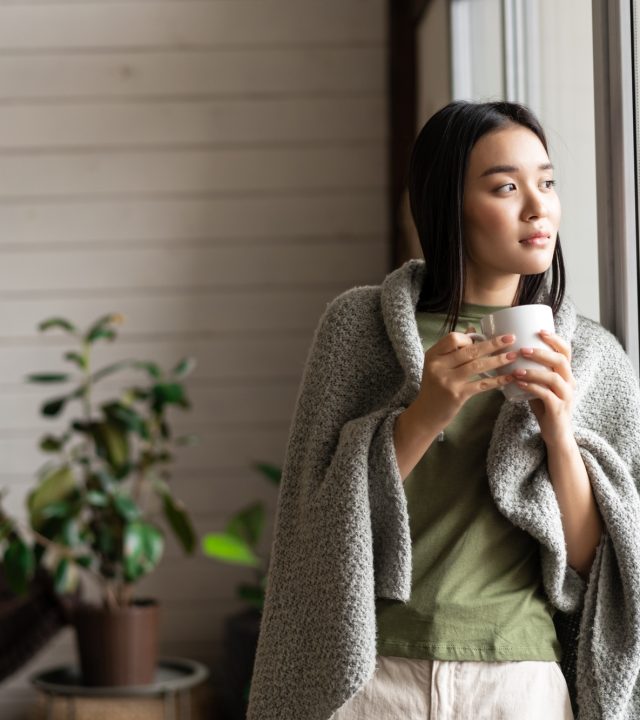 Beautiful asian woman wrapped in blanket, leaning on window and looking outside, drinking hot tea